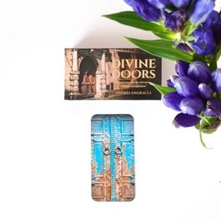 Book titled 'Divine Doors' by Andrea Engracia with a door illustration, next to a small model of a door and purple flowers on a white background.