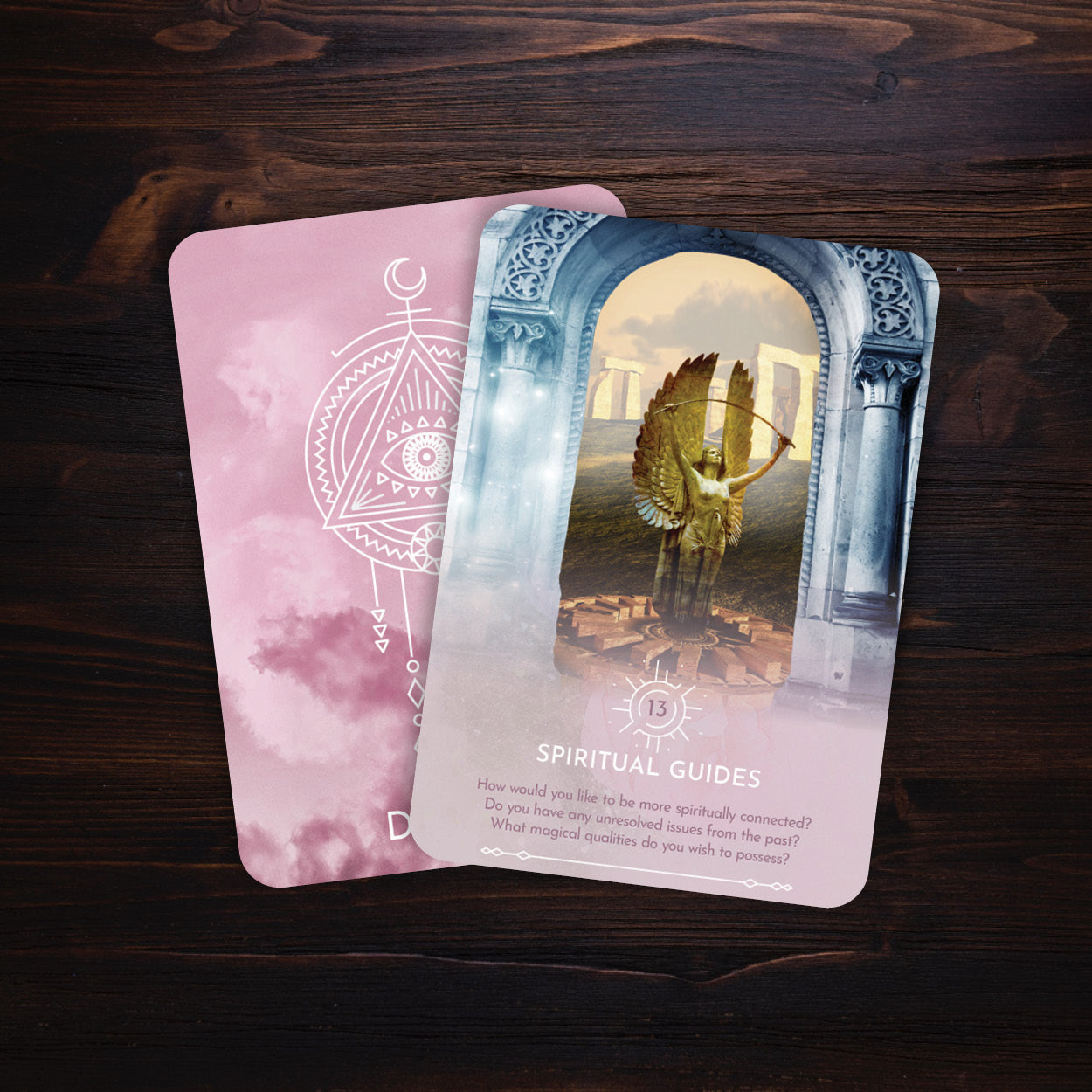 Two tarot cards on a wooden surface, one pink with a geometric design and the other with an angelic scene.